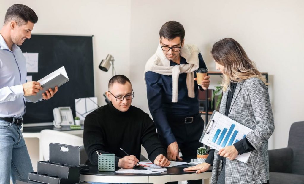Ryhmä taloushallinnon asiantuntijoita pöydän ympärillä. Kuvituskuva havainnollistaa, miten sosiaalinen oppiminen on keskeinen osa taloushallinnon osaamisen kehittämistä.